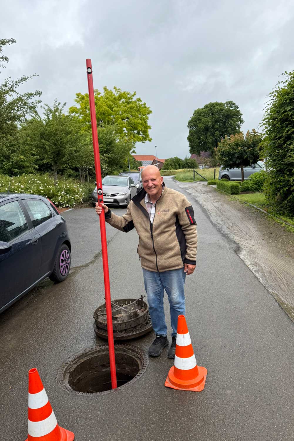 Vermesser hält eine rote Messlatte in einen geöffneten Kanalschacht, der mit Warnkegeln abgesichert ist.